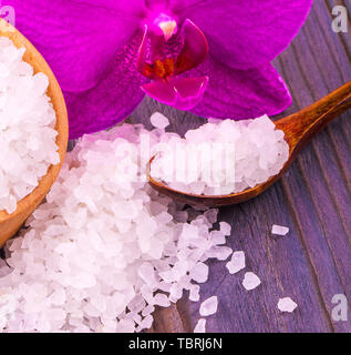 Das weiße Bad Salz in eine hölzerne Schale mit einem Löffel auf einen hölzernen Tisch mit einer Orchidee Stockfoto