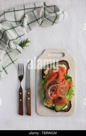 Big leckere Smorrebrod mit Lachs, Grün vegatbles, Zwiebeln, Kapern, Gurken, Käse, Pfeffer und Roggenbrot Festlegung auf weißem Handtuch auf Holzbrett. Messer und Gabel mit Holzgriffen sind in der Nähe. Stockfoto