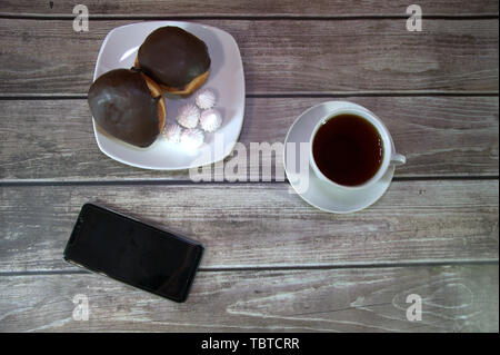 Eine Platte mit Donut und Marshmallow, einer Tasse Tee und einem Smartphone liegen auf einem Holztisch. Stockfoto