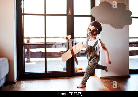 Ein Kleinkind Junge mit Karton Flugzeug drinnen zu Hause spielen, fliegen Konzept.