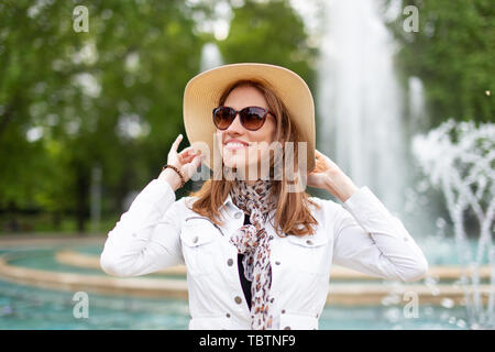 Glückliche junge stilvolle Frau in Sonnenbrille Holding hat im Park mit Springbrunnen Stockfoto