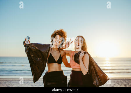 Freundliche weibliche Probanden mit Müllsack Gestik Sieg unterzeichnen. Frau Surfer am Strand mit Müllsäcken während des Sonnenuntergangs. Stockfoto