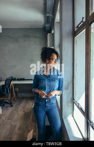 Junge Frau, die in Büro- und schaut aus dem Fenster. Inhaber kleiner Unternehmen in Casuals stehend durch die Fenster im Büro. Stockfoto