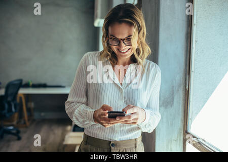 Geschäftsfrau, die mit Ihrem Smart Phone im Büro. Weibliche Unternehmer an Ihr Mobiltelefon suchen und lächelnd. Lesen von SMS-Nachrichten. Stockfoto