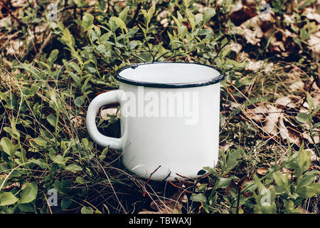 In der Nähe von Metall Becher auf Waldboden unter Heidelbeer Sträuchern und Eichenlaub. Tee, Kaffee im Freien Zeit. Mockup von weißen Emaille Tasse, Lifestyle entspannen, t Stockfoto