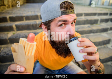 Trinken Sie etwas anders. Kaukasische hipster Trinken Essen trinken mit Hot Dog. Man nimmt einen Schluck frischen Kaffee trinken im verfügbaren cup Bärtigen. Ernährung Essen und Trinken. Stockfoto