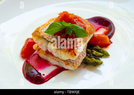 Nahaufnahme einer Pfanne gebratene gegrillter Red Snapper Fischfilet mit Spargel, rote Beete Sauce, Cherry San Marzano Tomaten und Basilikum auf einem weißen Teller natürliche Stockfoto