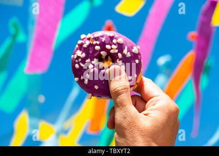 Eine appetitliche Donut, beschichtet mit einem violetten Zuckerguss und weiß besprüht, in der Hand des jungen Mannes gegen den Himmel, wo viele Fahnen der verschiedenen colo Stockfoto