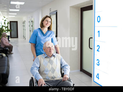 Lächelnd Krankenschwester Unterstützung älterer Mann im Rollstuhl im Krankenhaus Flur Stockfoto