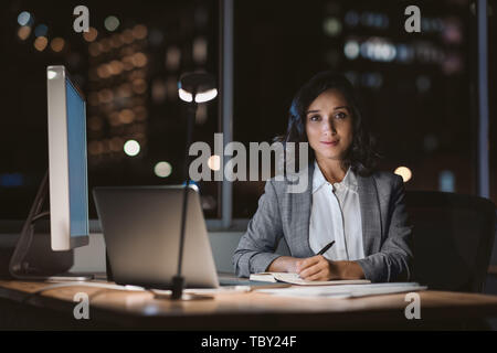Lächelnd geschäftsfrau an ihrem Schreibtisch sitzen in einem dunklen Büro Stockfoto