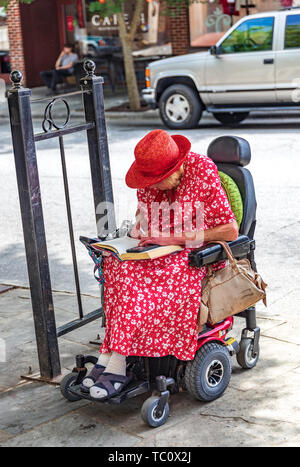ASHEVILLE, NC, USA -5/31/19: Eine behinderte, ältere Frau im roten Kleid und Red Hat, sitzt im Rollstuhl, ein Buch lesen und Rauchen. Stockfoto