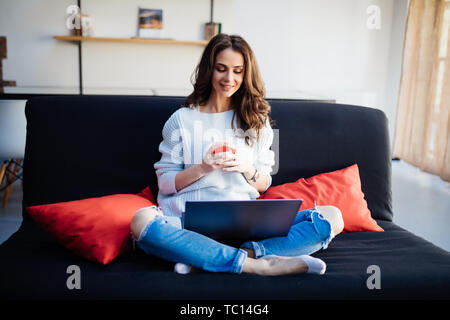 Lächelnd schöne Brünette auf dem Sofa entspannen und mit Ihrem Laptop im Wohnzimmer Stockfoto