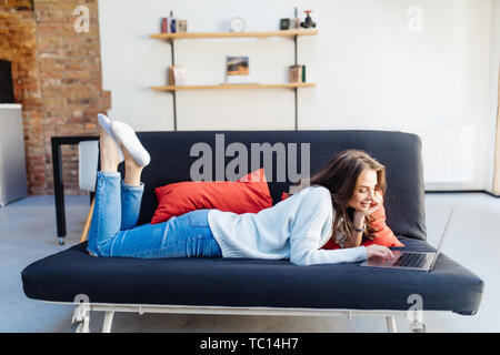 Frau liegt auf dem Sofa mit einem Laptop in ihrem Wohnzimmer Stockfoto