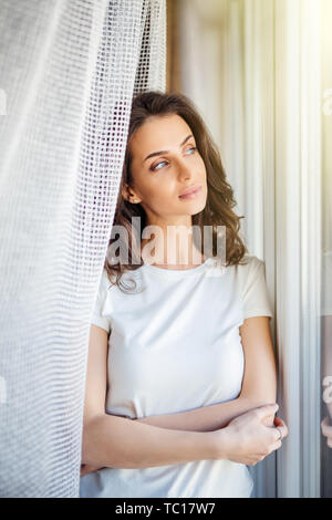 Seitenansicht eines einzigen nachdenkliche Frau stehend nach draußen durch ein Fenster und Vorhänge aus einem Wohnzimmer zu Hause öffnen Stockfoto