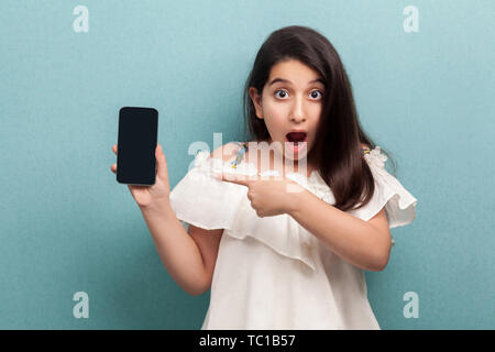 Portrait von schockiert Schöne jugendlicher Mädchen in weißem Kleid, die hielten Telefon und zeigen mit dem Finger auf Display mit geöffnetem Mund, bei Camera suchen Stockfoto