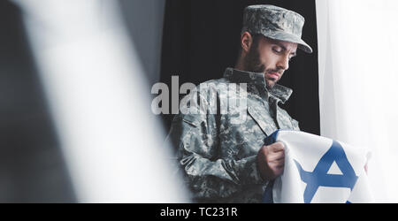 Selektiver Fokus der nachdenkliche militärischen Mann in Uniform holding Israel Nationalflagge, während man durch die Fenster Stockfoto