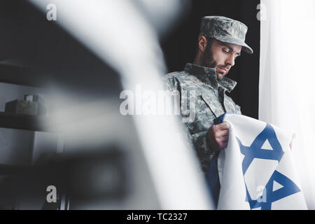 Selektiver Fokus der nachdenkliche bärtigen Mann in Uniform holding Israel Nationalflagge, während man durch die Fenster Stockfoto