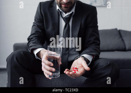 7/8-Ansicht der Mann im schwarzen Anzug hält Glas Wasser und eine Handvoll Pillen Stockfoto