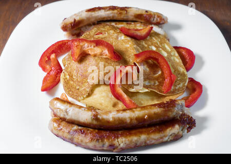 Gebratene Würstchen, Pfannkuchen und roter Pfeffer liegen auf einem weißen Teller. Holz- Hintergrund. Stockfoto