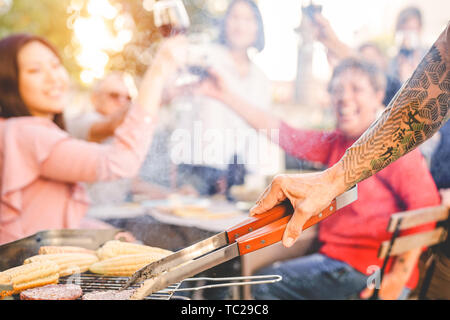 Tätowiert älterer Mann grillen Hamburger für seine Familie an Barbecue Dinner Party - Close up männliche Hand Kochen mit Grill im Garten Stockfoto