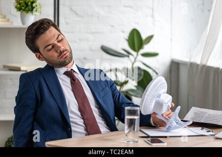 Stattliche erschöpft Unternehmer leiden unter Hitze, während am Arbeitsplatz vor Blasen elektrischer Ventilator sitzend Stockfoto