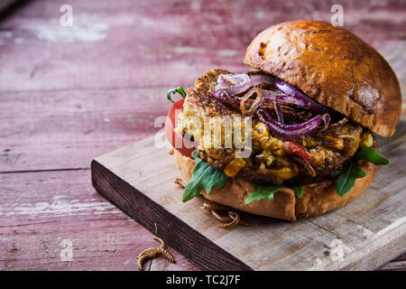 In der Nähe auf einem gebratenen mealworm Insekt Burger auf einem frischen cruty Brötchen mit Tomaten, Zwiebeln und Rucola Zutaten auf einem urigen Schneidebrett für Menü adv serviert. Stockfoto