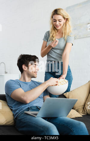 Gerne blonde Mädchen, dass Schüssel mit Popcorn in der Nähe von fröhlichen Mann sitzt mit Laptop auf dem Sofa Stockfoto