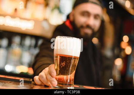 Bärtige und lächelnd Barkeeper gibt Bier. Cool und mutig Bar. Ein glückliches Barkeeper hält ein Bier in der Hand. Becher schäumenden Getränk. Stockfoto