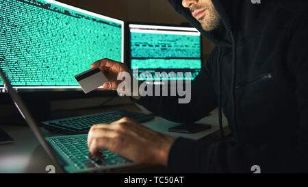 Diebstahl von Geld. Seitenansicht der junge Bartgeier Hacker in Schwarz hoodie mit mehreren Computern und Holding Kreditkarte beim Sitzen in dunklen Zimmer. Der binäre Code. Cyber Attack. Cyber Security Stockfoto