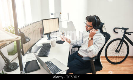 Sprechen über business. Seitenansicht der junge Bartgeier Händler im Headset holding Bericht und im Gespräch mit Kunden beim Sitzen vor Bildschirmen im Büro. Geschäftskonzept. Handel Konzept. Call Center Stockfoto