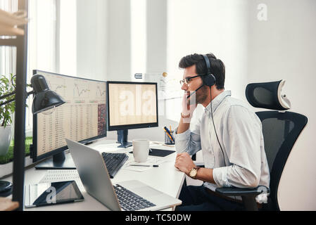 Immer verfügbar. Seitenansicht der junge Bartgeier Händler im Headset sprechen mit Kunden und den Blick auf den Bildschirm des Monitors mit dem Handel von Diagrammen und finanzielle Daten, während in seinem modernen Büro zu sitzen. Geschäftskonzept. Handel Konzept. Kommunikationskonzept Stockfoto