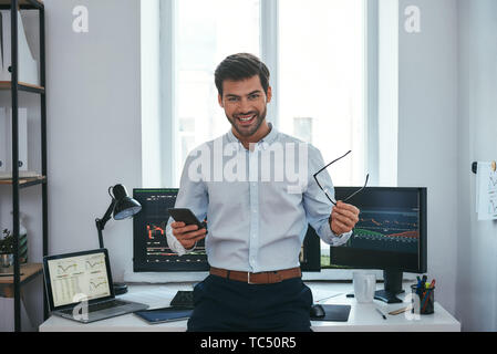 Tolle Happy junge Unternehmer oder Unternehmer in formale Holding Brillen und Smartphone und lächelnd, während vor den Bildschirmen mit den Charts im Büro. Forex Markt. Geschäftskonzept. Investitionen Konzept Stockfoto