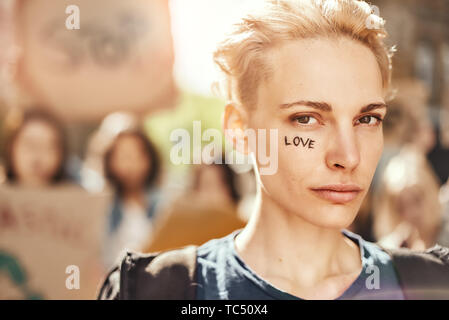 Macht Liebe, nicht Krieg. Nahaufnahme Foto, junge blonde Frau mit dem Wort Liebe auf ihrem Gesicht stand vor der Aktivistinnen auf der Straße während Protest März geschrieben. Menschen demonstriert. Protest Konzept. Revolution Konzept Stockfoto