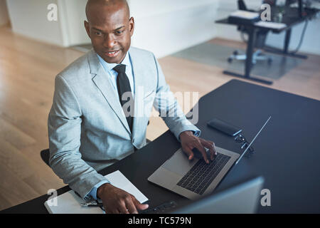 Jungen afrikanischen amerikanischen Geschäftsmann, Lächeln, während sie online arbeiten mit einem Laptop an seinem Schreibtisch in einem modernen Büro Stockfoto