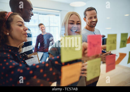 Team verschiedener Designer bei einem Brainstorming mit Haftnotizen auf eine Glaswand lächelnd in einem Büro Stockfoto