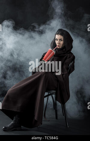 Frau in den Tod Kostüm sitzt auf einem Stuhl und trinken Trinken auf Schwarz Stockfoto