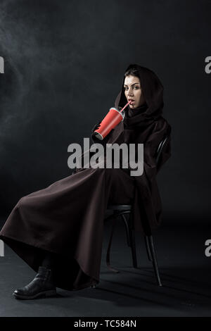 Frau in den Tod Kostüm sitzt auf einem Stuhl und trinken Trinken auf Schwarz Stockfoto