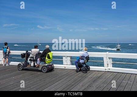 Behinderte Frau im Rollstuhl und Behinderte im Duo zwei Person Mobilität scooter Boote beobachten auf See von Jetty Stockfoto