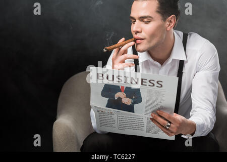 Mann sitzt im Sessel lesen Zeitung und Rauchen Zigarre auf Schwarz mit Rauch Stockfoto