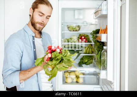 Junge vegan Mann wählen, was zu kochen und dabei frisches Gemüse aus dem Kühlschrank zu Hause Stockfoto