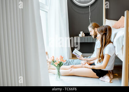 Junge Paar genießt die Morgen sitzen zusammen mit Buch und Kaffee in der Nähe des Bettes vor dem Fenster zu Hause Stockfoto