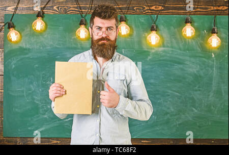 Bärtige hipster hält Buch, schwarzes Brett für den Hintergrund. Lehrer in Brillen empfiehlt Buch, Daumen hoch. Empfehlung Konzept. Mann mit Bart und Schnurrbart auf ernstes Gesicht vor der Tafel Stockfoto