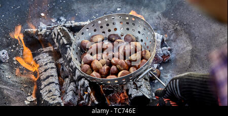 Panorama Banner eines Mannes Röstung frische Kastanien in einem Metall Röster über glühende Kohlen und einen Grill Feuer im Freien in einer ersten Person POV Stockfoto