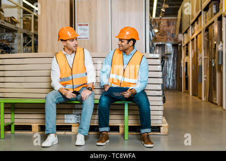 Lächelnd multikulturellen Arbeitnehmer in Sicherheitswesten und Helme ruhen auf der Werkbank im Lager Stockfoto