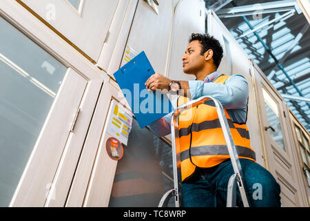 Stattlich, konzentrierte indischen Lagerarbeiter stehend auf Leiter und die Zwischenablage Stockfoto