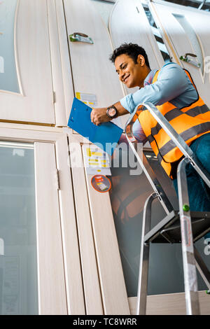 Lächelnd indischen Lagerarbeiter stehend auf Leiter und die Zwischenablage Stockfoto