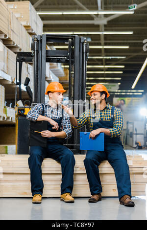 Zwei lächelnde multikulturellen Arbeitnehmer mit den Klemmbrettern Sprechen während der Sitzung auf Sperrholz Stockfoto