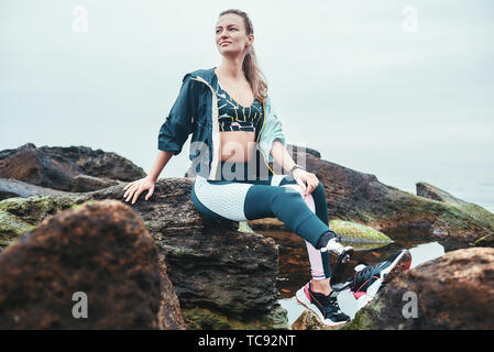 Ausruhen nach Training. Gerne behinderte Athleten Frau in Sportswear mit beinprothese saß auf dem Stein und denken über das Leben. Sport Konzept. Motivation. Behinderte Sportler. Stockfoto