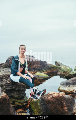 Glücklich zu leben. Positive behinderte Athleten Frau in Sportswear mit beinprothese saß auf dem Stein und Kamera mit Lächeln. Sport Konzept. Motivation. Behinderte Sportler. Stockfoto