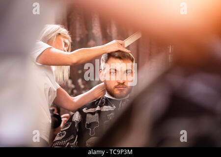 Der Master schneidet die Haare und Bart eines Mannes in einem Friseursalon, Ein Friseur ist ein Haarschnitt für einen jungen Mann. Schönheit Konzept, Self Care. Stockfoto